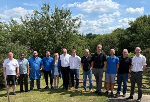 RVL team photo taken outdoors in the sunshine, with blue sky and white clouds with green trees behind the people being photographed. Staff members are standing in a curved line, with MP Mike Martin in the centre. The visit took place to show the visitor how RVL is involved in UK electronics design and manufacturing.
